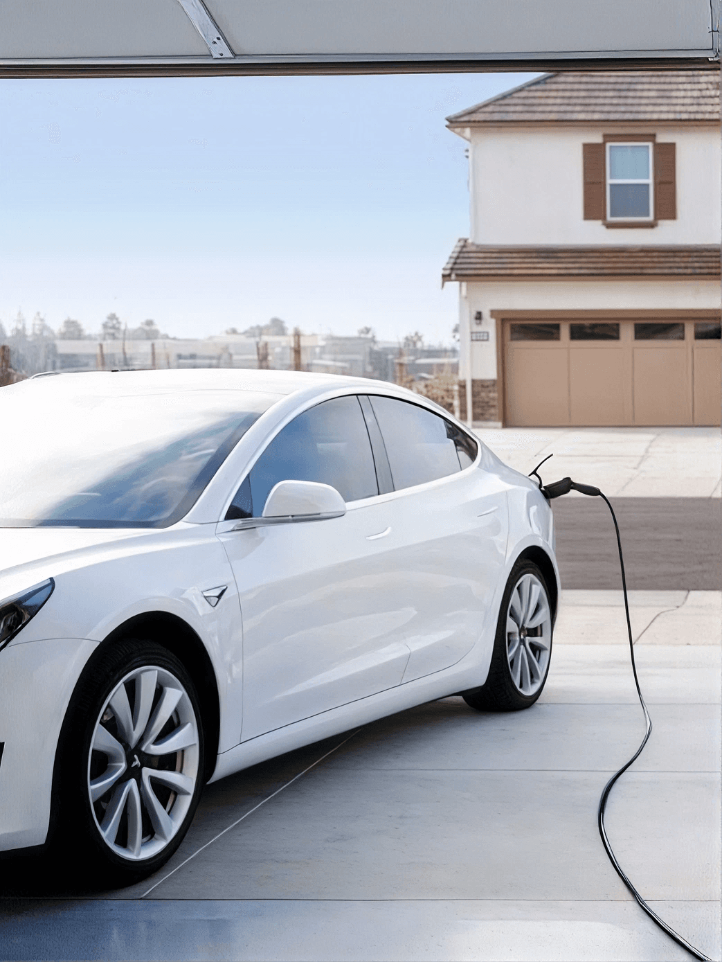 A white electric car charges in a driveway, connected by a charging cable. A modern house with a two-car garage and blue sky are in the background.