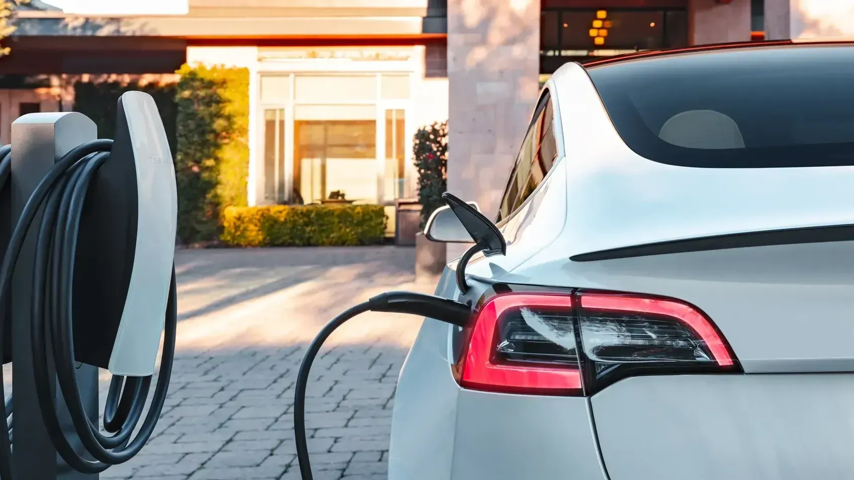 A white electric car is parked in a residential driveway, plugged into a charging station. The scene conveys modernity and eco-friendliness.