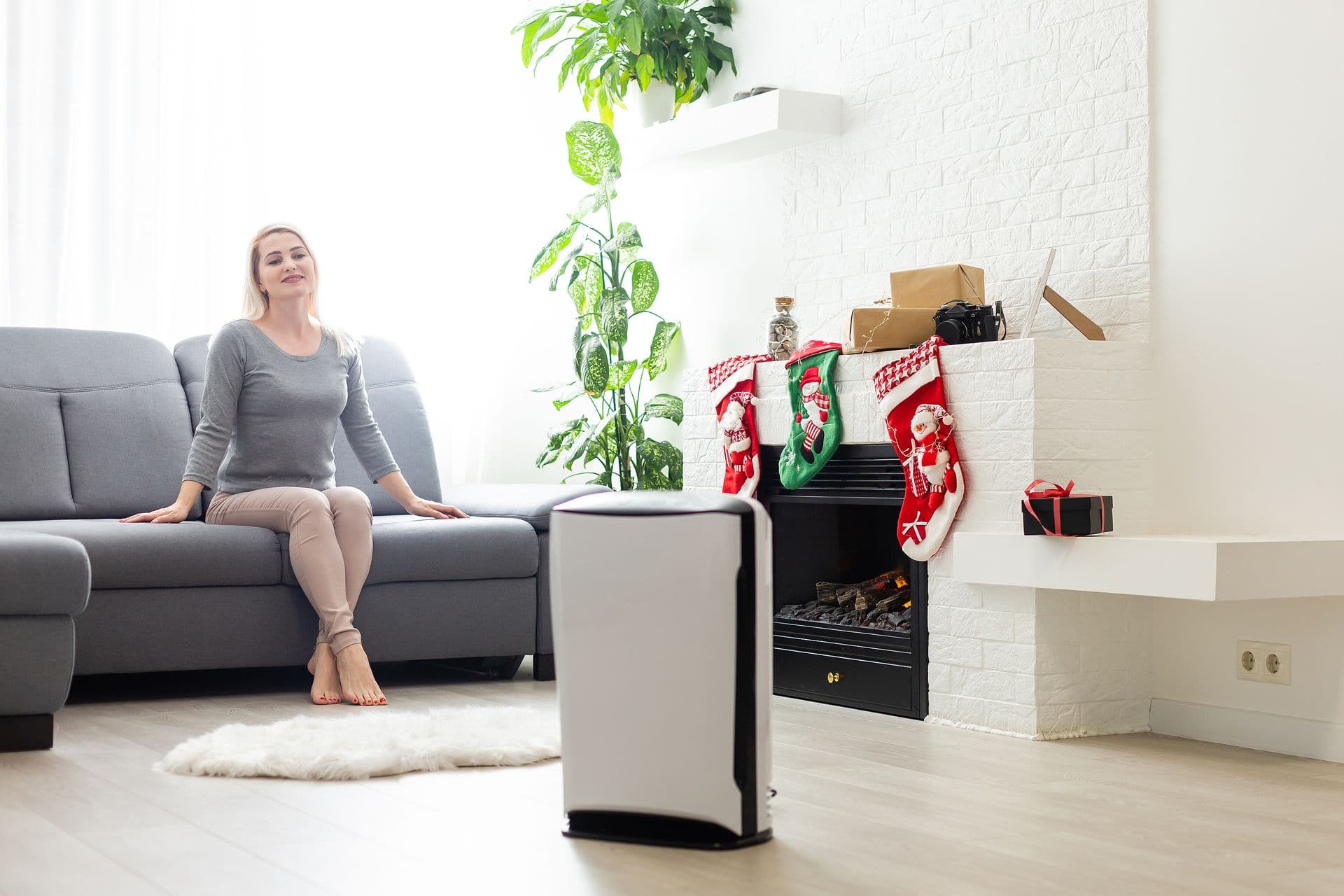 A woman sits relaxed on a gray sofa in a bright, modern living room. A fireplace is decorated with Christmas stockings, gifts, and a potted plant.