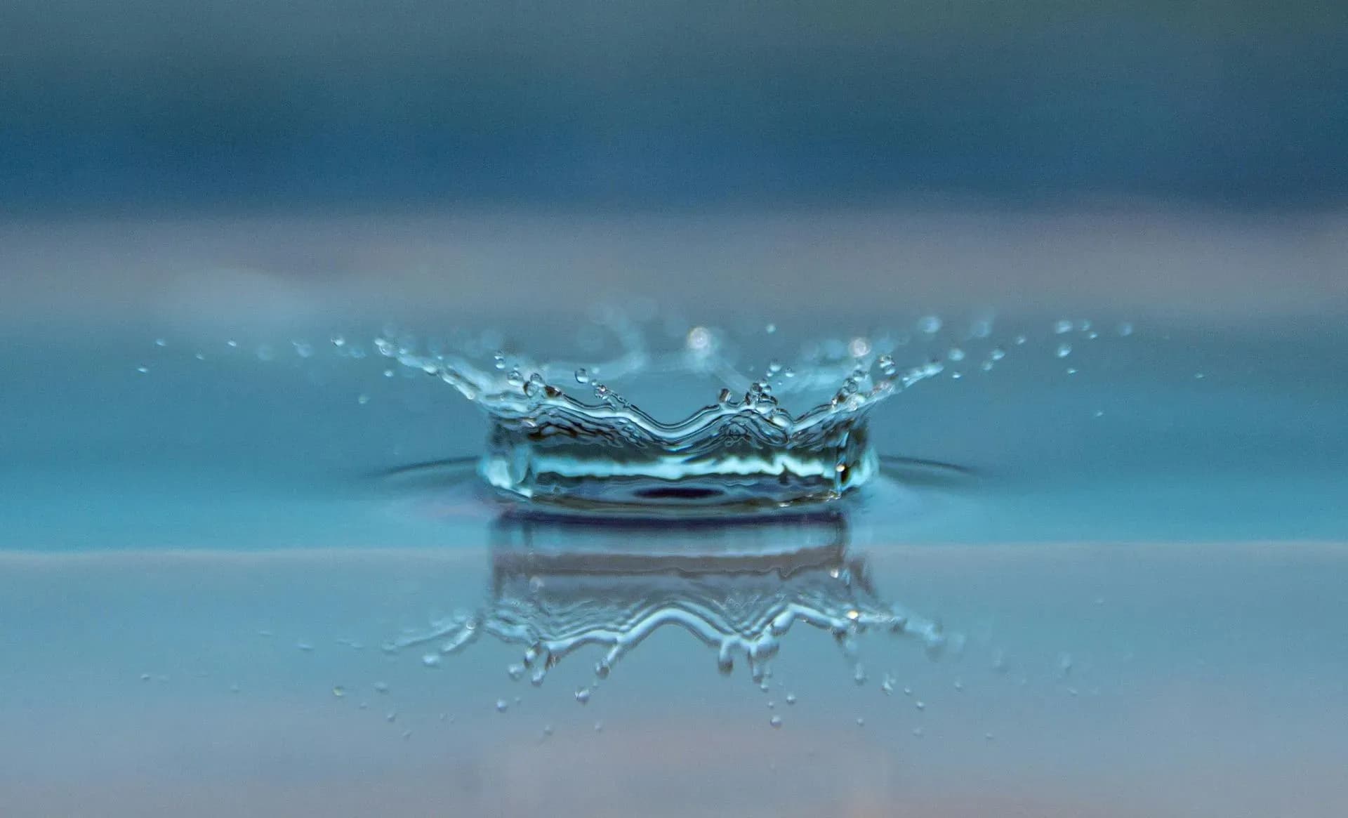 A water droplet creates a ripple and splash, forming a crown shape on a smooth blue surface. The scene is calm and serene, conveying tranquility.
