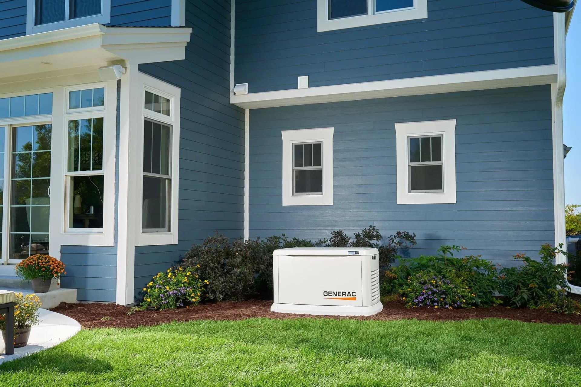 A blue house exterior featuring white trim, three large windows, and a Generac generator beside bushes and flowers. The yard is lush and green.