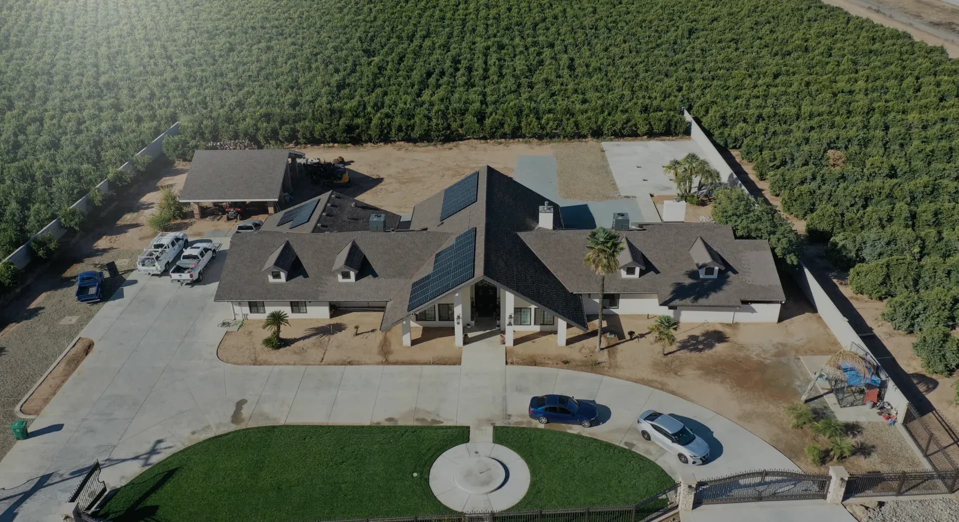 Arial view of a large single-story house with a brown roof and solar panels, surrounded by a lush green orchard. A driveway circles the front, where cars are parked.