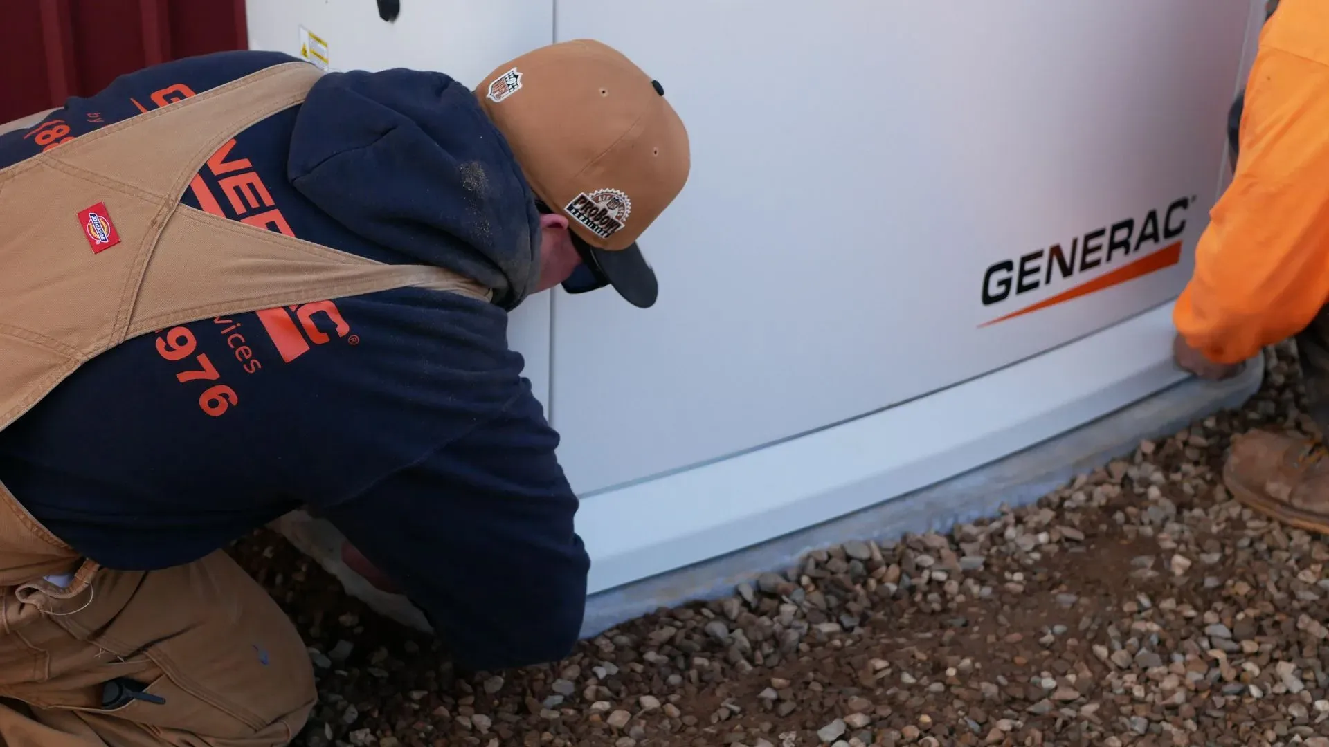 Two workers in outdoor gear are installing a Generac generator on a gravel surface. They are focused and working together in a cooperative manner.