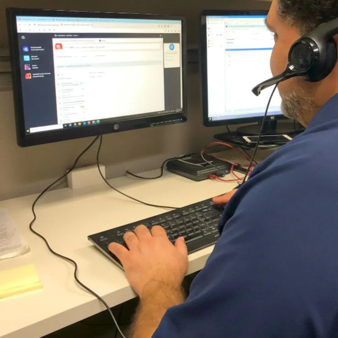 A person wearing a headset works at a desk with dual monitors displaying software applications. They type on a keyboard, focused on the task.