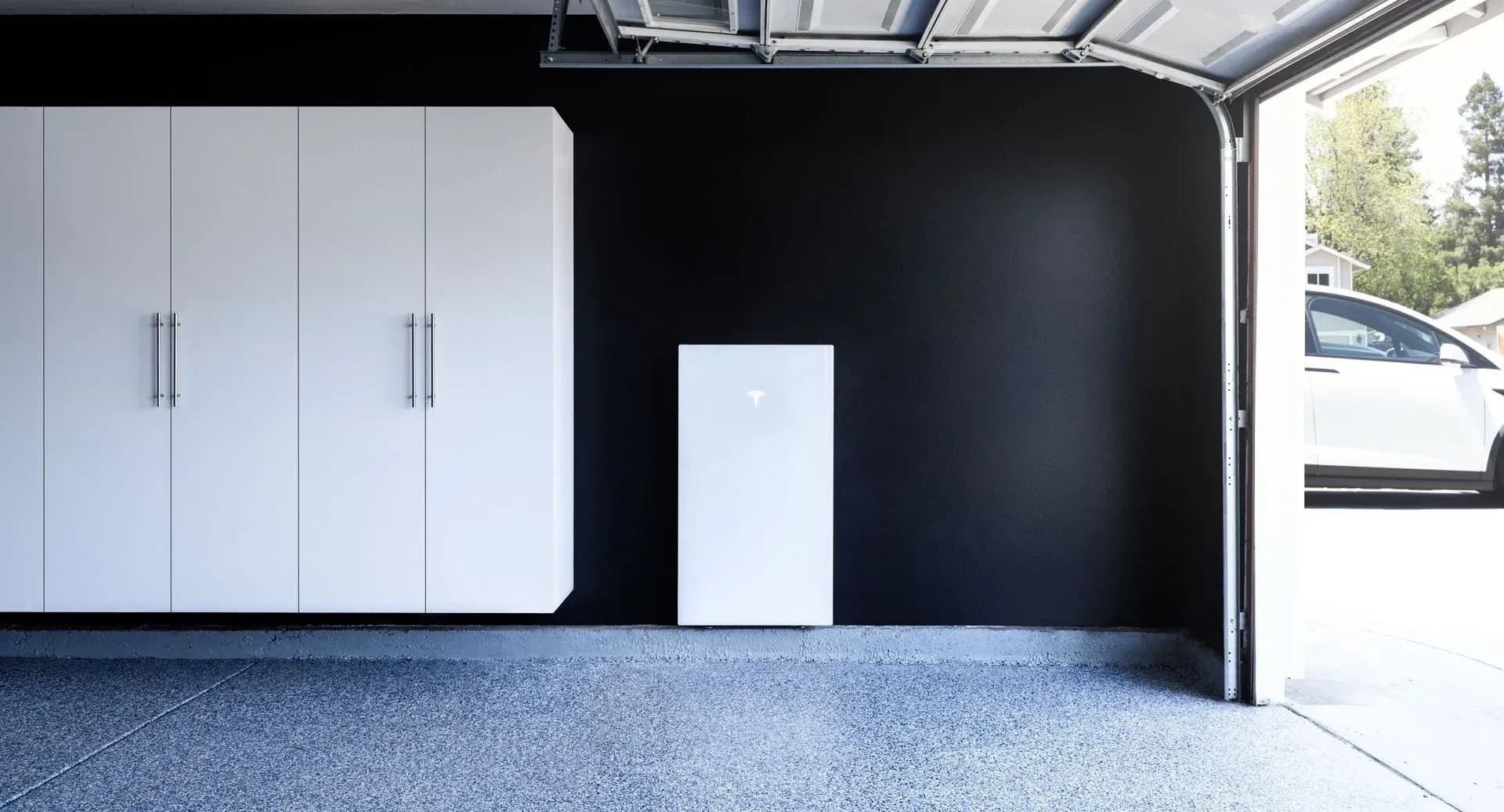A modern garage with sleek white cabinets on a black wall, featuring a Tesla Powerwall. The garage floor is polished gray, and a white car is visible outside.